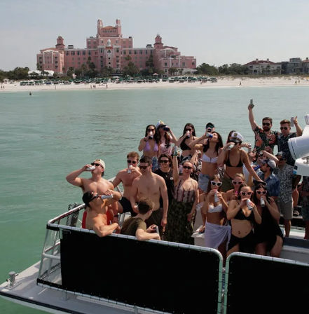 Sunny boat party with a crowd in swimsuits drinking on turquoise water, white-sand beach and large pink beachfront hotel in the background.