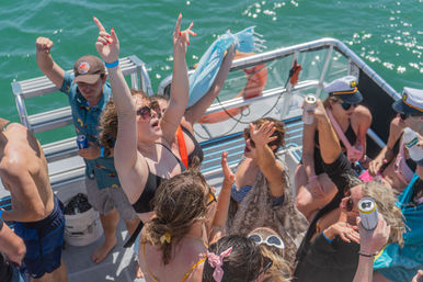 Energetic group partying on a sunny pontoon boat, wearing swimsuits and captain hats, dancing and raising cans over green summer water