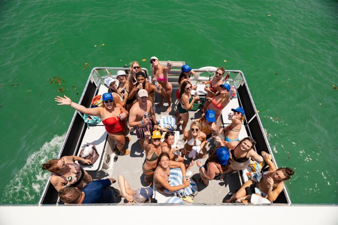 Aerial view of a lively summer pontoon boat party with a group of friends in swimsuits waving and holding drinks on emerald-green water.