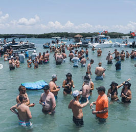 Crowded sandbar boat party in shallow clear water, dozens of people in swimwear socializing near anchored pontoons and boats on a sunny summer day