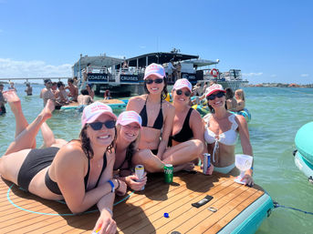 Five women in bikinis wearing pink caps and sunglasses pose on a floating wood platform at a sunny boat party in turquoise coastal water with drinks and inflatables nearby.