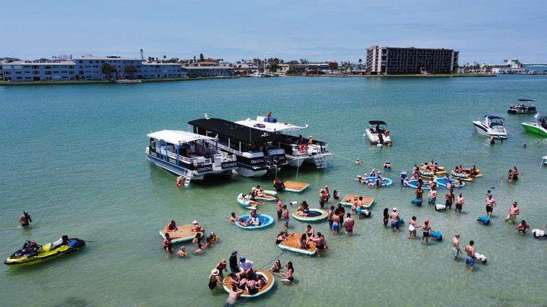 Sunny coastal bay with a summer beach party: groups of people on floating rafts and inner tubes near anchored party boats and jet skis, waterfront condos on the horizon.