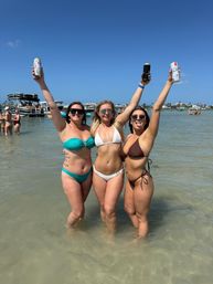 Three friends in colorful bikinis standing waist-deep in clear shallow water on a sunny beach, arms raised cheering with canned drinks; pontoon boats and a coastal shoreline under a bright blue sky in the background.