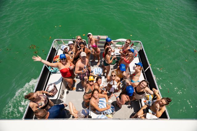 Aerial view of a lively pontoon boat party with young adults in swimsuits cheering, holding drinks and sunbathing on a sunny summer day over clear green coastal water