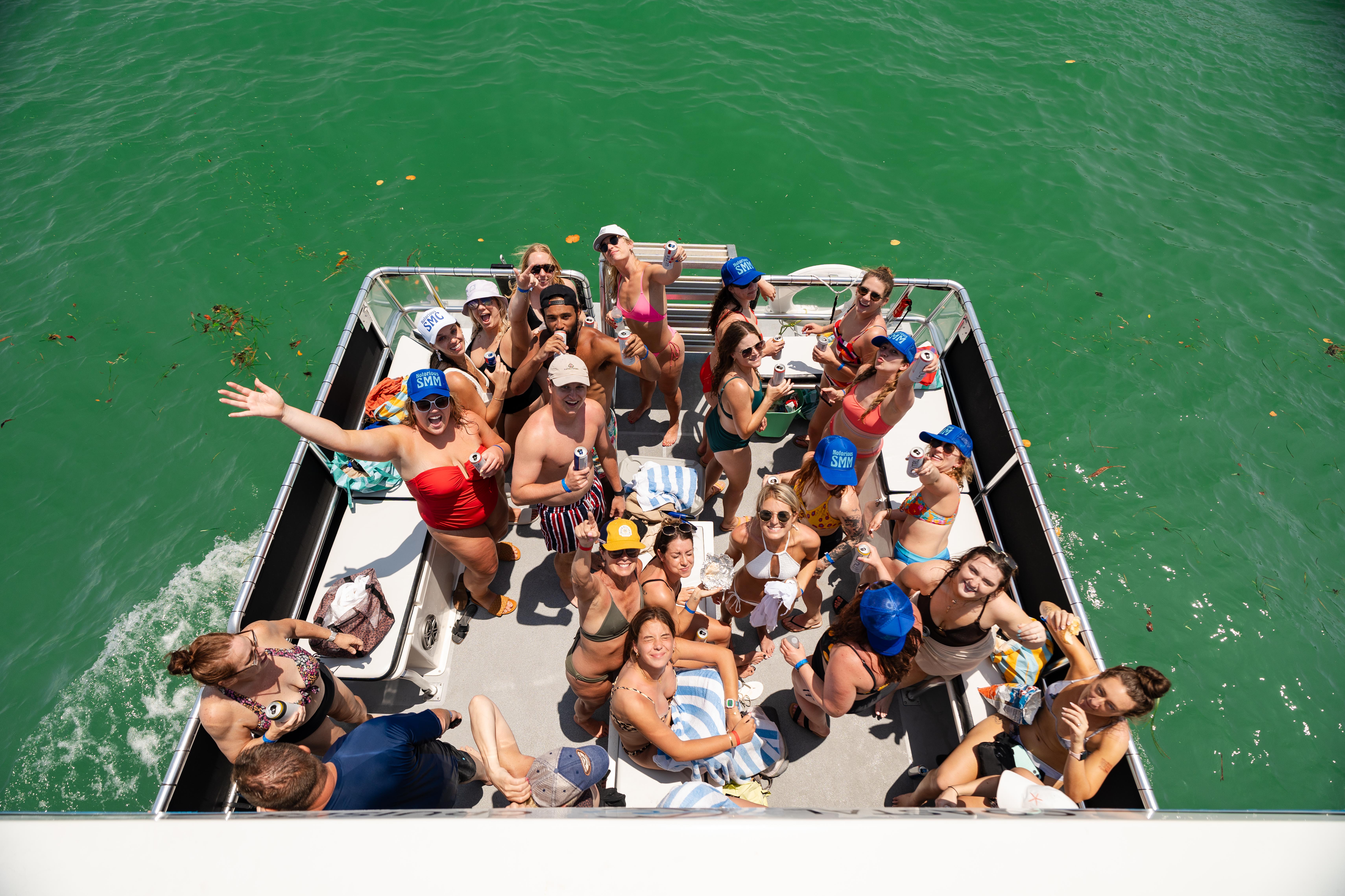 Summer pontoon boat party with a large group of young adults in colorful swimwear waving and raising drinks on a sunny day, surrounded by clear emerald-green water.
