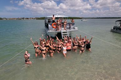 Group of people in bikinis at a beach boat party, wading in shallow turquoise water near an anchored party boat on a sunny day