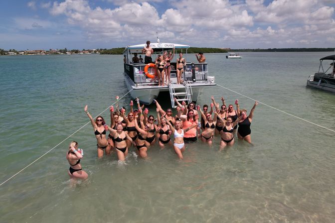 Group of people in bikinis at a beach boat party, wading in shallow turquoise water near an anchored party boat on a sunny day