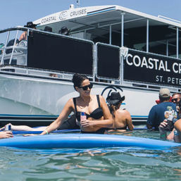 Woman in black bikini and sunglasses sitting on a blue paddleboard with a canned drink, floating near a large white passenger boat and other sunbathers on a sunny coastal day.