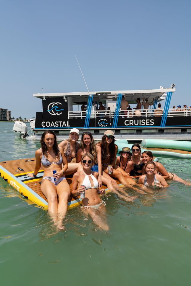 Group of friends in bikinis lounging on a large inflatable dock in clear coastal water, holding drinks with a party cruise boat in the background under a bright blue sky — sunny beach-day vibe.