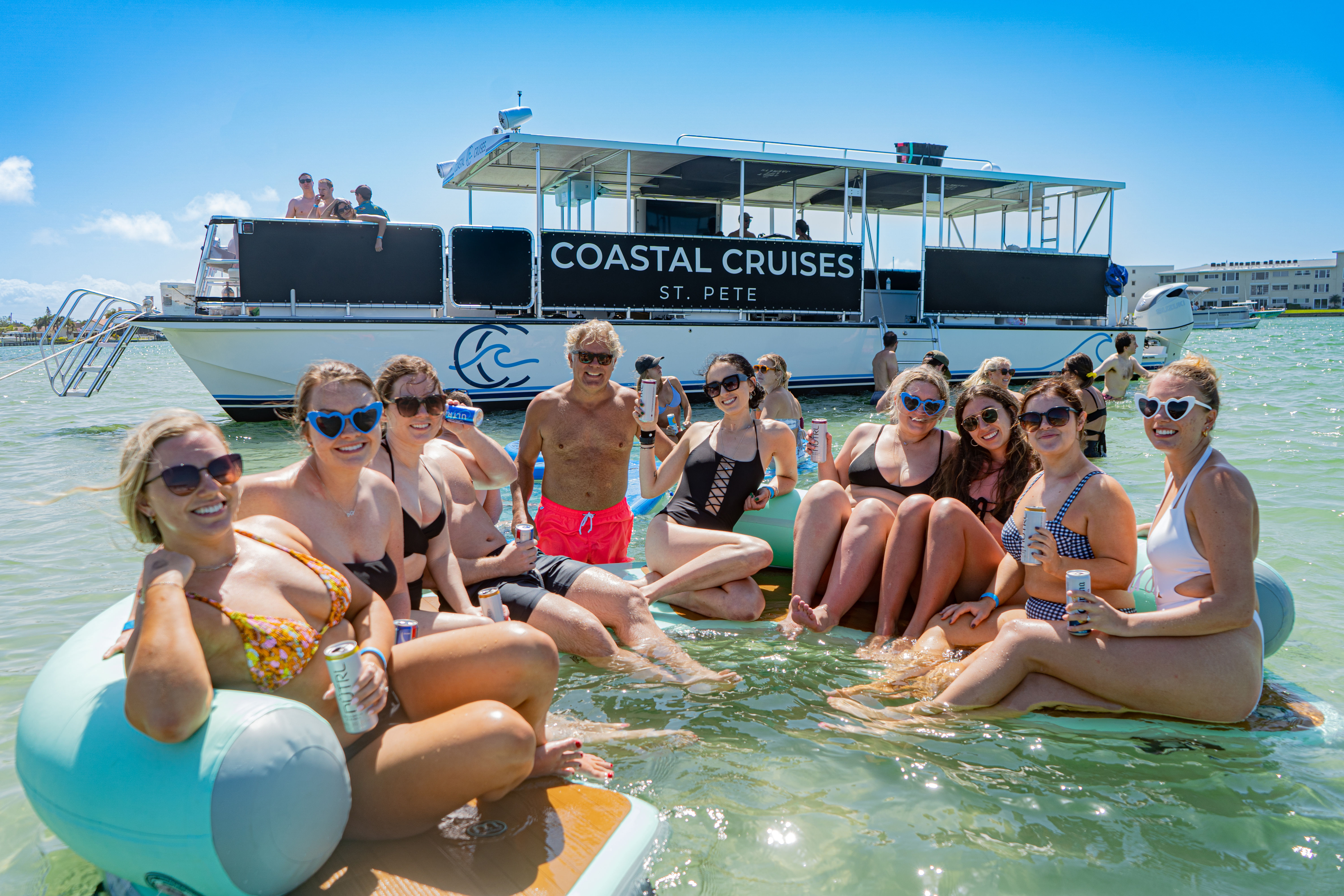 Group of friends lounging on inflatable tubes in shallow turquoise coastal water beside a large tour boat, smiling in swimsuits and holding canned drinks on a sunny day