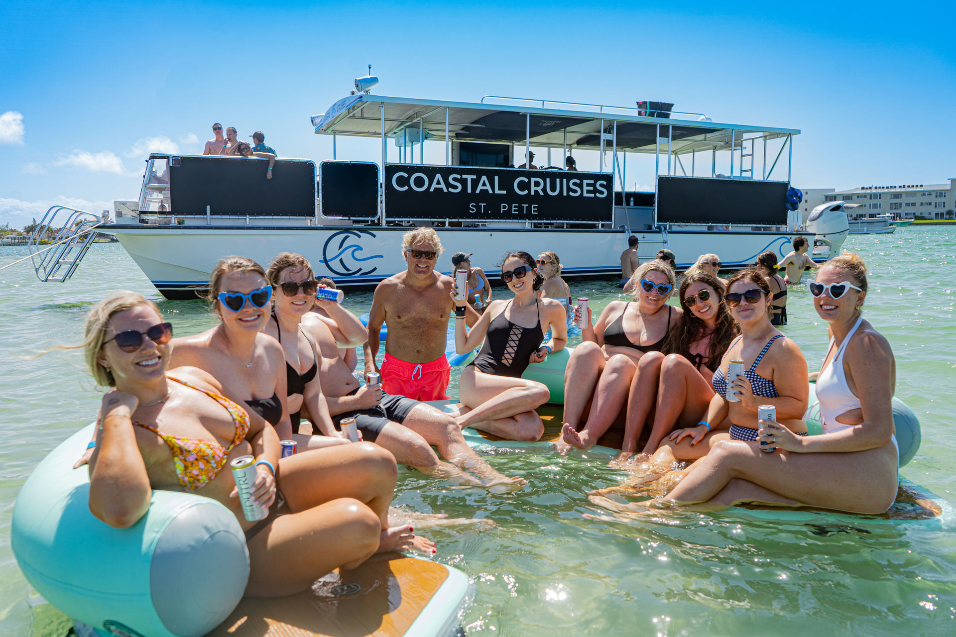 Smiling group of friends lounging on inflatable floats with drinks in shallow turquoise water by a party cruise boat off St. Pete, Florida.