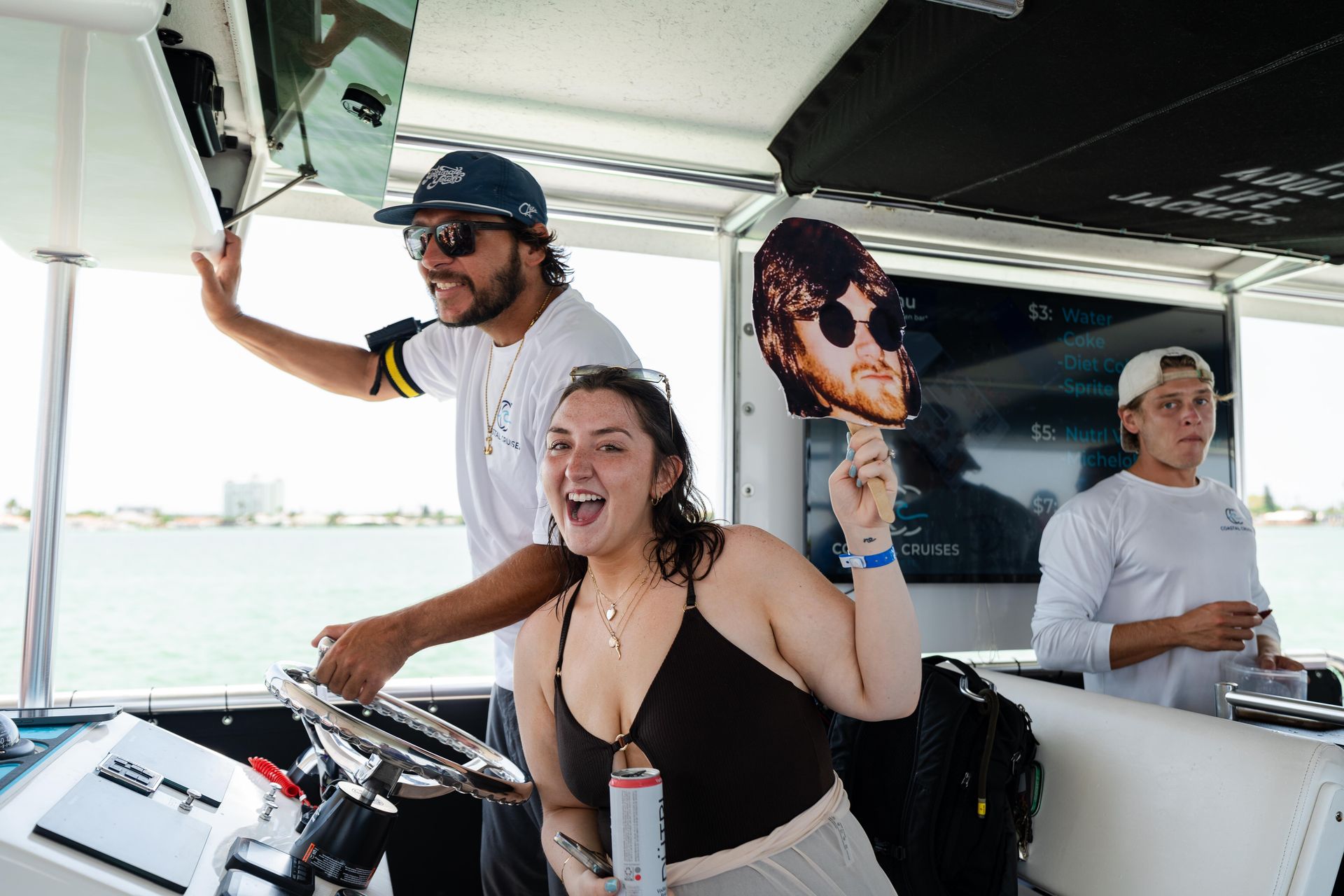 Smiling woman holding a cutout face and canned drink at the helm of a sunny daytime boat cruise, crew members steering with coastal skyline in background