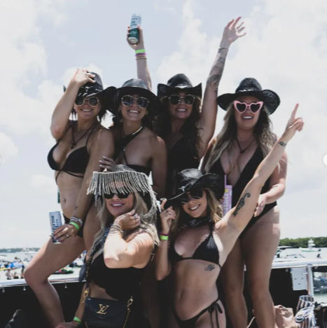 Six women in black bikinis and cowboy hats posing on a boat during a sunny lake boat party, wearing sunglasses, holding drinks and raising their arms in celebration.