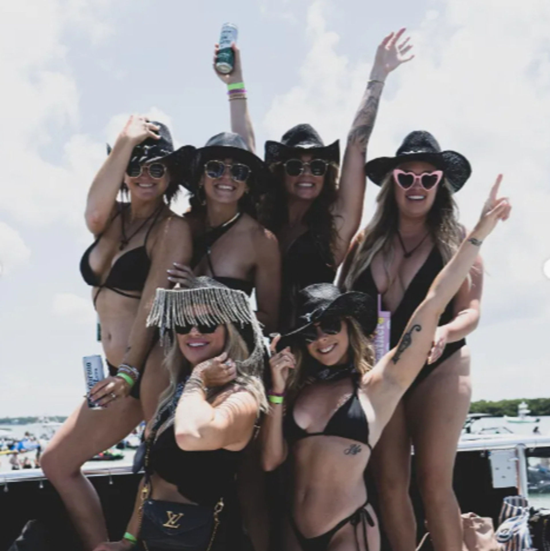 Six women in black bikinis and cowboy hats posing on a boat during a sunny lake boat party, wearing sunglasses, holding drinks and raising their arms in celebration.