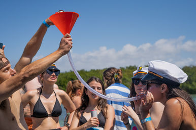 People on a sunny boat party passing drinks through an orange funnel and clear tube, wearing swimwear and captain hats with a mangrove-lined coast and blue sky in the background