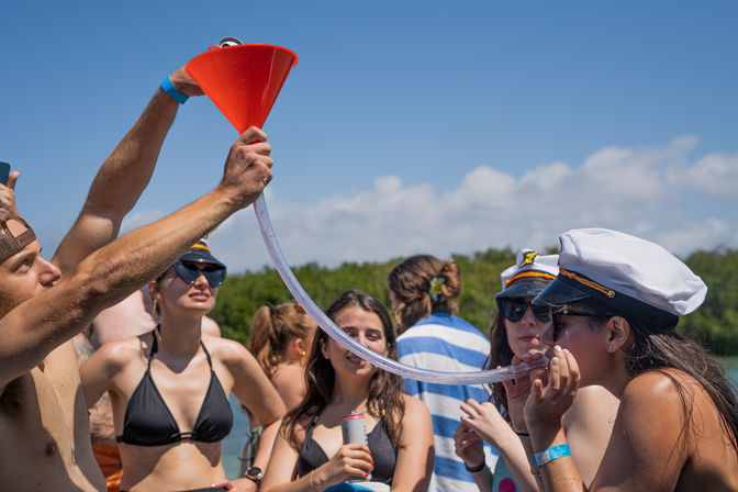 People on a sunny boat party passing drinks through an orange funnel and clear tube, wearing swimwear and captain hats with a mangrove-lined coast and blue sky in the background