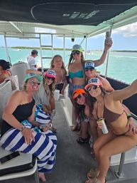 Smiling group of friends in bikinis and colorful trucker hats enjoying canned drinks on a motorboat over turquoise tropical water with low islands and sunny sky.