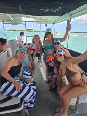 Smiling group of friends in bikinis and colorful trucker hats enjoying canned drinks on a motorboat over turquoise tropical water with low islands and sunny sky.