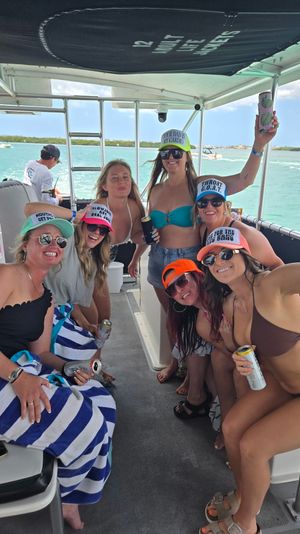 Smiling group of friends in bikinis and colorful trucker hats enjoying canned drinks on a motorboat over turquoise tropical water with low islands and sunny sky.