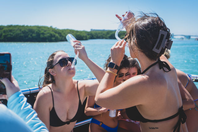 Group of friends in bikinis on a boat passing drinks and toasting on a sunny day over turquoise water and a tree-lined shoreline