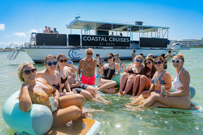 Group of friends in swimsuits lounging on inflatable tubes in shallow turquoise water off St. Pete, Florida, holding drinks and posing in front of a docked party cruise boat on a sunny day.
