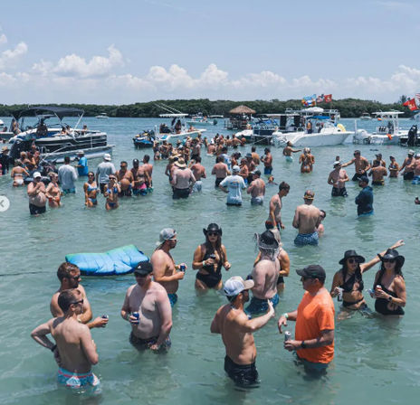 Crowded sandbar boat party with dozens of people wading in shallow turquoise water, chatting and holding drinks amid anchored boats and pontoons under a sunny blue sky.