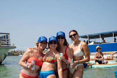 Four smiling women in colorful swimsuits wearing blue "Notorious SMM" caps, holding canned drinks while waist-deep in clear coastal water beside boats and paddleboards on a sunny summer day.