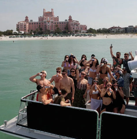 Boat party of young adults holding drinks and posing on turquoise water off a sandy beach with a large pink beachfront resort in the background