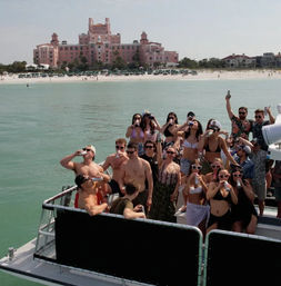 Group of people in swimsuits drinking and posing on a party boat in turquoise water off a sandy beach with a large pink seaside hotel in the background on a sunny day.
