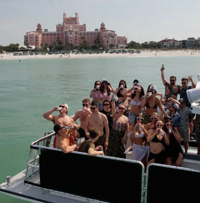 Boat party of young adults holding drinks and posing on turquoise water off a sandy beach with a large pink beachfront resort in the background