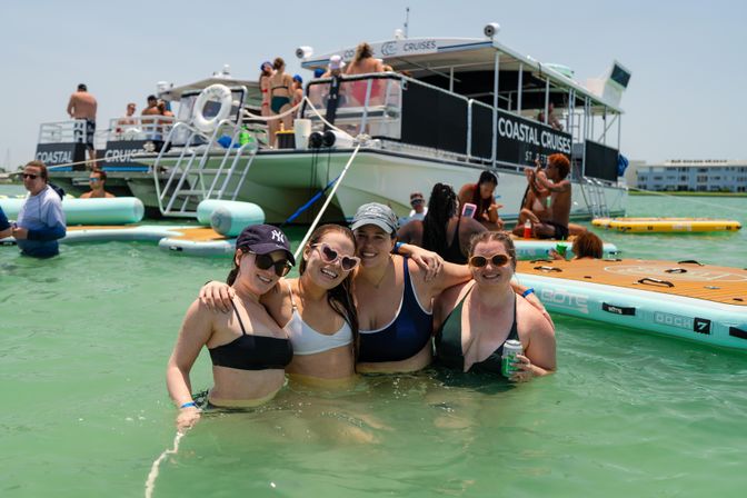 Four smiling women in swimsuits posing in shallow green coastal waters beside a party boat and inflatable platforms on a sunny day