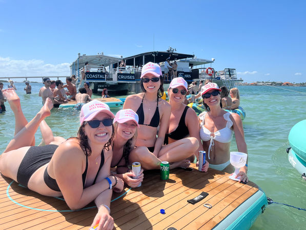 Five women in bikinis wearing pink caps and sunglasses pose on a floating wood platform at a sunny boat party in turquoise coastal water with drinks and inflatables nearby.