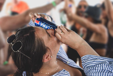 Person in striped shirt with sunglasses on their head tilting a blue can to their mouth amid a blurred crowd at a lively summer party