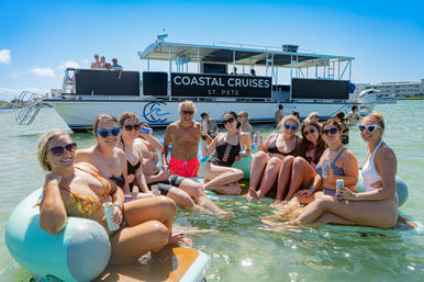 Smiling group of friends lounging on inflatable floats with drinks in shallow turquoise water by a party cruise boat off St. Pete, Florida.