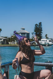 Person wearing a purple birthday party hat and black bikini sipping from a champagne flute while holding a canned drink on a boat, sunny coastal marina with waterfront homes and boats in the background.