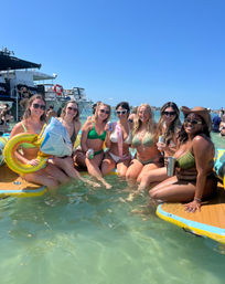 Group of friends in bikinis relaxing on paddle floats in clear shallow coastal water at a sunny boat and beach party, holding drinks and inflatable props (one wearing a bachelorette sash).