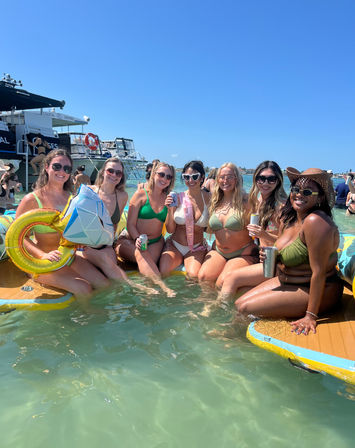 Group of friends in bikinis relaxing on paddle floats in clear shallow coastal water at a sunny boat and beach party, holding drinks and inflatable props (one wearing a bachelorette sash).