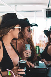 Three women in black swimsuits and cowboy hats wearing sunglasses, smiling and holding canned drinks aboard a sunny beach party boat.