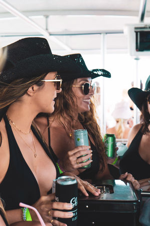 Three women in black swimsuits and cowboy hats wearing sunglasses, smiling and holding canned drinks aboard a sunny beach party boat.