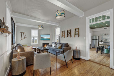 Bright open-plan coastal-style living room with hardwood floors, gray sofa and armchairs, colorful pendant lamps, wall-mounted TV above a lit fireplace, decorative wooden ladder shelves, and a view into a stainless-steel kitchen.