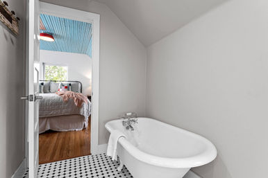 Freestanding clawfoot bathtub on black-and-white hex tile with gray walls, open door offering a peek into a cozy bedroom featuring hardwood floors and a blue beadboard ceiling.