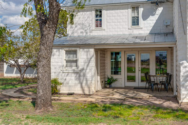 Sunlit white stone house patio with metal roof, glass double doors, a small outdoor bistro table and chairs, potted plants, and a large shade tree on a grassy yard