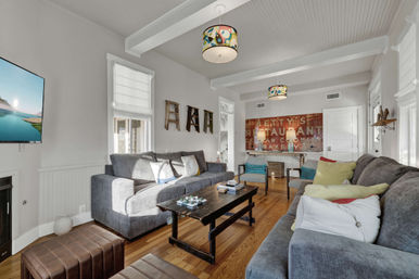 Sunlit living room with gray sofas and colorful throw pillows, hardwood floors, white beadboard walls, patterned pendant lights, black wooden coffee table, wall-mounted TV and vintage-style wall décor