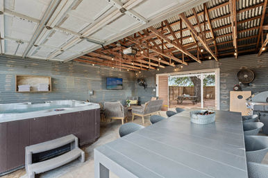 Rustic covered backyard patio with hot tub, long outdoor dining table, lounge seating, string lights and exposed wooden beams opening to a tree-shaded yard.