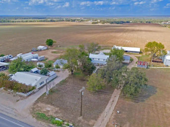 Aerial view of a country farmhouse and barns amid sprawling fallow fields, dirt drive and utility pole, framed by mature trees under a bright blue, cloud-dotted sky.