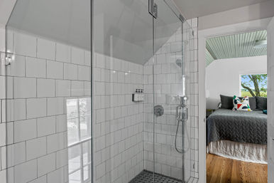 Glass-enclosed walk-in shower with white subway tile, sloped ceiling, black pebble floor and chrome fixtures, opening to a bright bedroom with gray quilted bed, colorful accent pillow, hardwood floor and a window view of green trees.