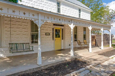 Sunny rural farmhouse front porch with white stone exterior, yellow door, hanging wooden porch swing, vintage metal chairs, and decorative star-cut trim.