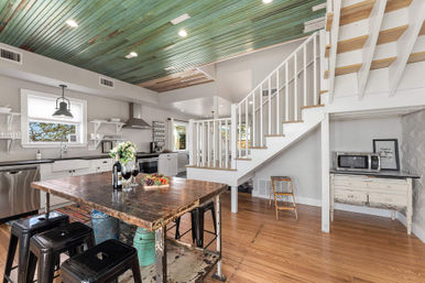 Bright open-concept coastal farmhouse kitchen with distressed wooden island and black metal stools, green painted beadboard ceiling, white staircase, hardwood floors, stainless appliances, and fresh flowers on the table