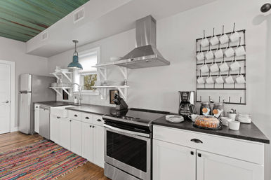 Bright modern farmhouse kitchen with white cabinets, stainless steel appliances, farmhouse sink, open shelving, wall mug rack, coffee station, and colorful woven rug.