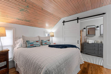 Cozy attic bedroom with sloped painted wood ceiling, quilted gray bed and patterned accent pillows, bedside lamps and sliding barn door leading to an en-suite bathroom with gray vanity and black-and-white tiled floor.